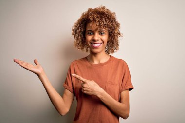 Kıvırcık saçlı güzel Afro-Amerikalı kadın beyaz arka plan üzerine günlük tişörtler giyip kameraya hayret ediyor ve gülümsüyor..