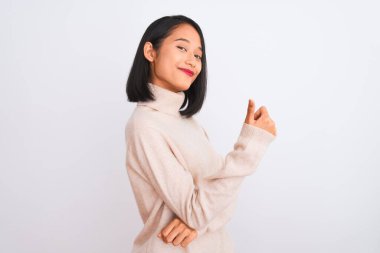 Balıkçı yaka kazak giyen genç bir kadın soyutlanmış beyaz arka planda dikiliyor gururlu, gülümseyen, baş parmak kaldıran bir jest yapıyor.