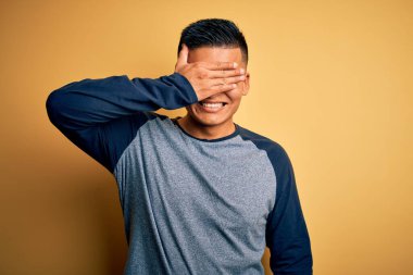 Sarı arka planda dikilen gündelik tişört giyen genç, yakışıklı, Latin bir adam. Gülümsüyor ve elleri yüzünde, şaşırtmak için gözleri kapatıyordu. Kör kavram.