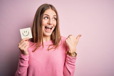 Genç, güzel, kızıl saçlı bir kadın elinde gülümseyen bir kâğıt, gülümseyen bir yüz ifadesiyle işaret eden ve baş parmağıyla gösteren bir emoji mesajı tutuyor.