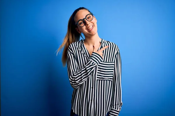 Beautiful blonde woman with blue eyes wearing striped shirt and glasses over blue background cheerful with a smile of face pointing with hand and finger up to the side with happy and natural expression on face