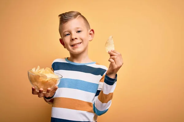 若い白人の子供食べるUnheathyポテトチップス上の黄色の背景に幸せな顔立ちと笑顔で自信を持って笑顔を示す歯 — ストック写真