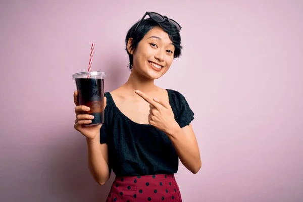 年轻美丽的中国女人喝可乐时 用稻草覆盖粉红的背景 手指手画脚 非常高兴 — 图库照片