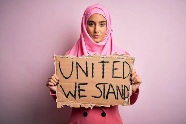 ピンクのイスラム教徒のヒジャーブを身に着けている若い活動家の女性が団結してバナーを保持私たちは真剣に考えてスマート顔に自信を持って表現でメッセージを立つ — ストック写真