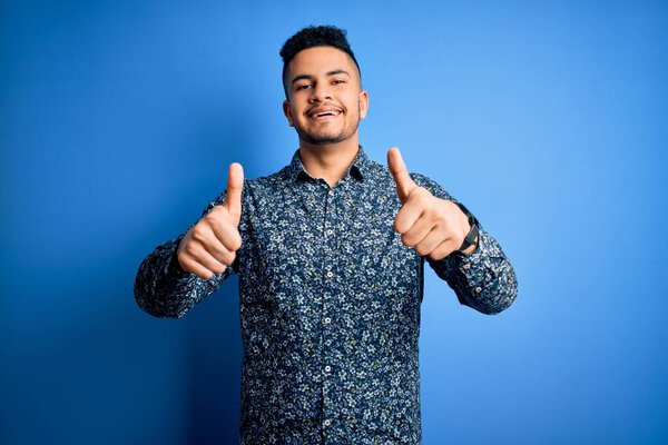 Young handsome man wearing casual shirt standing over isolated blue background success sign doing positive gesture with hand, thumbs up smiling and happy. Cheerful expression and winner gesture.