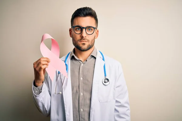 Young doctor man wearing stethoscope holding pink ribbon about cancer over isolated background with a confident expression on smart face thinking serious