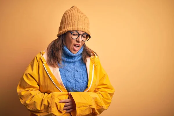 Middle age woman wearing yellow raincoat and winter hat over isolated background with hand on stomach because nausea, painful disease feeling unwell. Ache concept.
