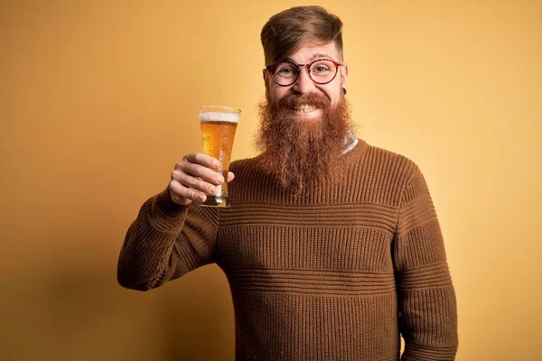 Irish redhead man with beard drinking a glass of refreshing beer over yellow background with a happy face standing and smiling with a confident smile showing teeth