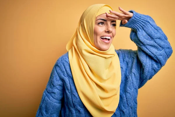 Young beautiful brunette muslim woman wearing arab hijab over isolated yellow background very happy and smiling looking far away with hand over head. Searching concept.