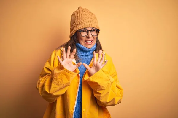Middle age woman wearing yellow raincoat and winter hat over isolated background disgusted expression, displeased and fearful doing disgust face because aversion reaction. With hands raised