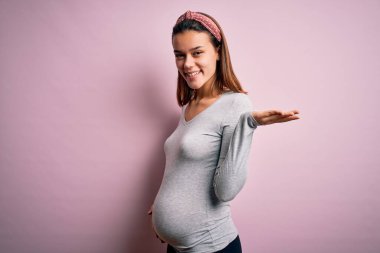 Genç, güzel, hamile bir kız pembe arka planda bebek bekliyor. Gülümsüyor, avucunun içi gibi gülümsüyor ve kameraya bakıyor..