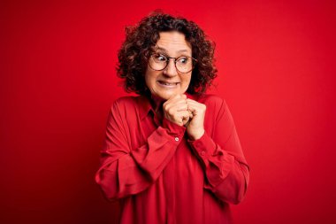 Orta yaşlı, kıvırcık saçlı, rahat gömlekli, kırmızı arka planda gözlüklü, gergin ve heyecanlı bir kadın.
