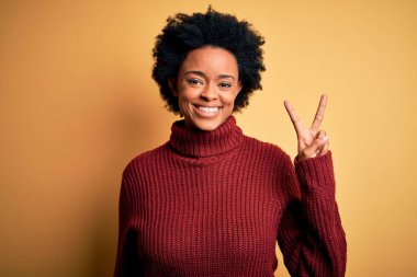 Kıvırcık saçlı, balıkçı yaka kazak giyen genç, güzel Afro-Amerikan kadın iki numarayı parmaklarıyla gösterirken kendinden emin ve mutlu gülümsüyor..