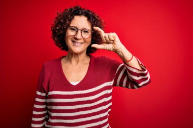 Orta yaşlı güzel kıvırcık saçlı kadın kırmızı arka planda sıradan çizgili süveter giyiyor gülümsüyor ve kendine güvenen el hareketleriyle küçük beden işareti yapıyor parmaklarıyla ve kamerayla bakıyor. Ölçü kavramı.