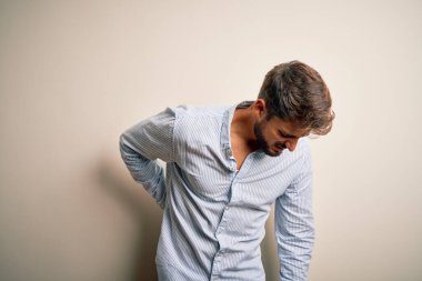 Sakallı, çizgili tişörtlü genç bir adam beyaz arka planda duruyor sırt ağrısı çekiyor, elle dokunuyor, kas ağrısı çekiyor.
