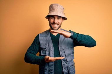 Tatilde sakallı yakışıklı bir turist sarı arka plan şapkası takıyor. Büyük ve büyük boylarda işaret eden el kol hareketleri yapıyor. Ölçü sembolü. Kameraya bakıp gülümsüyor. Ölçüm konsepti.