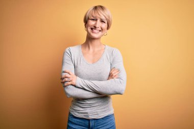 Sarışın, modern kısa saçlı, sarı arka planda duran mutlu yüzlü, kollarını kavuşturup kameraya bakan genç bir kadın. Pozitif kişi.