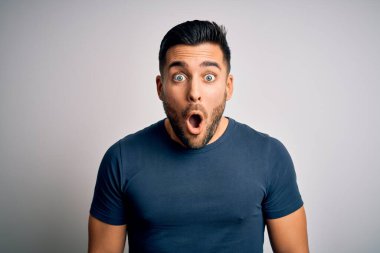 İzole edilmiş beyaz arka plan üzerinde duran sıradan tişört giyen genç yakışıklı adam korku, şaşkınlık ifadesi ve heyecanlı yüz ifadesiyle dehşete düşmüş ve şok olmuştu..