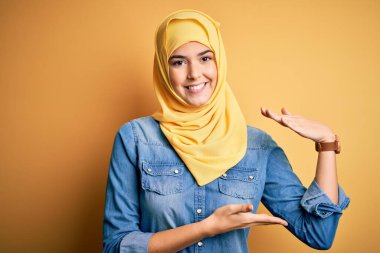İzole edilmiş sarı arka plan üzerinde duran Müslüman başörtüsü takan genç ve güzel bir kız. Ellerinde büyük ve büyük bir işaret, ölçü sembolü var. Kameraya bakıp gülümsüyor. Ölçüm konsepti.