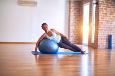 Orta yaşlı güzel sporcu kadın mutlu gülümsüyor. Spor salonunda yoga sınıfında spor topu kullanarak esneme hareketi.