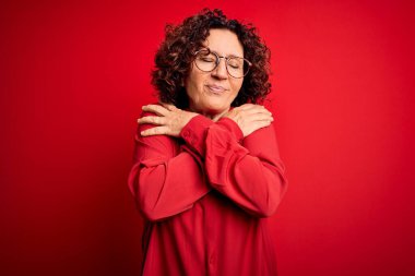 Orta yaşlı, kıvırcık saçlı, rahat gömlekli, kırmızı arka planda gözlüklü güzel bir kadın mutlu, pozitif ve kendinden emin bir şekilde kendine sarılıyor. Sevgi ve özgüven.