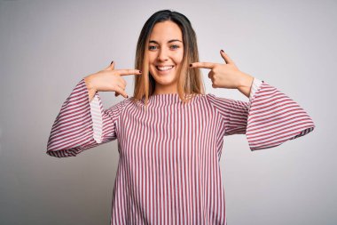 Mavi gözlü genç, güzel sarışın kadın beyaz arka planda ince tişörtler giyiyor neşeli gülüyor ve parmakları, dişleri ve ağzı ile işaret ediyor. Diş sağlığı konsepti.