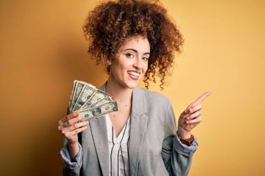 Kıvırcık saçlı, piercingli, dolarlar tutan genç ve güzel bir kadın. Elleri ve parmakları kenarı işaret ederek çok mutlu.