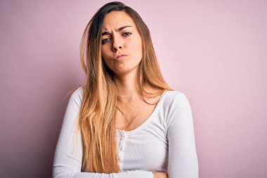 Pembe arka plan üzerinde beyaz tişört giyen, mavi gözlü genç, güzel sarışın kadın şüpheci ve gergin, yüzünde çapraz kollu tasvip etmeyen bir ifade var. Negatif kişi.