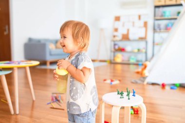 Sevimli çocuk yüzünde gülümsemeyle elinde şişeyle anaokulunda bir sürü oyuncağın etrafında duruyor.