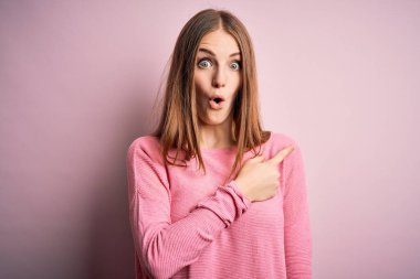 Genç, güzel, kızıl saçlı bir kadın pembe arka planda rahat bir süveter giyiyor. Parmakla işaret ediyor, ağzı açık, şaşkın bir ifade..