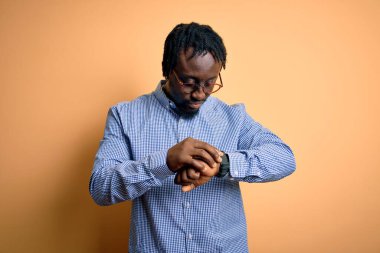 Sarı arka planda gömlek ve gözlük takan yakışıklı Afro-Amerikalı genç adam saati kontrol ediyor, rahat ve kendinden emin.