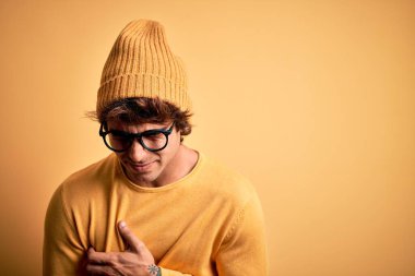 Genç, yakışıklı bir adam günlük tişört ve gözlük takıyor. El ve karın üstü izole edilmiş sarı bir arka plan. Çünkü mide bulantısı, acı verici bir hastalık. Ache kavramı.