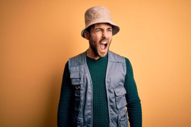 Tatilde sakallı yakışıklı bir turist sarı arka planda kaşif şapkası takıyor kızgın ve öfkeli çığlıklar atıyor, öfkeyle bağırıyor. Öfke ve agresif kavram.