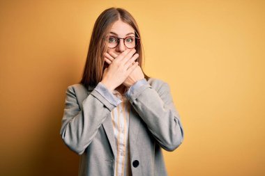 Sarı arka planda ceket ve gözlük takan güzel, kızıl saçlı bir kadın yanlışlıkla ağzı ellerle kapatmış. Gizli konsept.