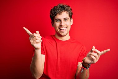 Kıvırcık saçlı, kırmızı arka planda sıradan tişörtler giyen, kendine güvenen, farklı yönleri gösteren genç, sarışın bir adam. Reklam için boşluk kopyala