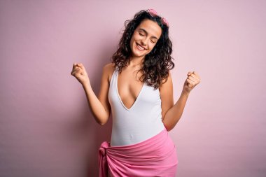 Tatilde kıvırcık saçlı güzel bir kadın pembe arka planda beyaz mayo giyerek çok mutlu ve heyecanlı bir şekilde kollarını kaldırıp, gülümseyerek ve başarı için çığlık atarak zafer hareketi yapıyor. Kutlama konsepti.