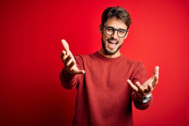 Sakallı, gözlüklü ve süveterli genç adam kırmızı arka planda dikiliyor. Gülümsüyor, yardım eli uzatıyor ve kabul ediyor..