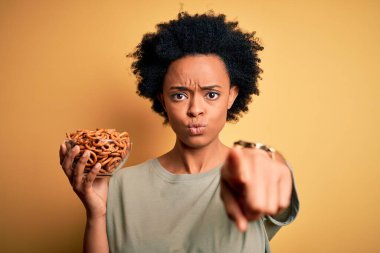 Afrika kökenli, kıvırcık saçlı, elinde fırında Alman krakerleri olan genç bir afro kadın kamerayı işaret ediyor ve sen, el işareti, ön taraftan pozitif ve kendinden emin bir jest.