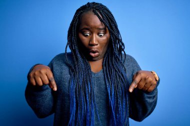 Afro-amerikan kadın örgü örüyor mavi arka planda günlük süveter giyiyor parmaklarını göstererek reklamını yapıyor, şaşırmış yüzünü ve ağzını açıyor.