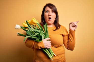Sarı arka planda romantik lale çiçekleri tutan güzel, iri yarı bir kadın. Parmakla işaret ederek şaşırmış, ağzı açık, şaşkın bir ifade..