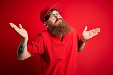 Gözlüklü ve kırmızı şapkalı genç teslimatçı soyutlanmış arka plan hakkında hiçbir şey bilmiyor ve elleri ve kolları havada şaşkın bir ifade sergiliyor. Şüphe kavramı.