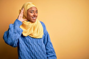 Genç Afrikalı Afro kadın, izole edilmiş sarı arka plan üzerine tesettür takmış, el pençe divan gülümseyerek dedikoduları ya da dedikoduları dinliyor. Sağırlık kavramı.