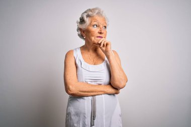 Son sınıf güzeli, gri saçlı, beyaz arka plan üzerine yazlık bir elbise giyen, elleri çenesinde soru soran, düşünceli bir kadın. Düşünceli bir yüzle gülümsüyor. Şüphe kavramı.