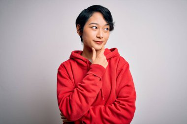 Genç, güzel Asyalı bir kız beyaz arka planda kapüşonlu bir kazak giyiyor. Elleri çenesinde soru sorarken, düşünceli bir ifade takınıyor. Düşünceli bir yüzle gülümsüyor. Şüphe kavramı.