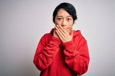 Beyaz arka planda kapüşonlu sıradan bir sweatshirt giyen genç güzel Asyalı kız ağzını yanlışlıkla ellerle kapatarak şok etti. Gizli konsept.