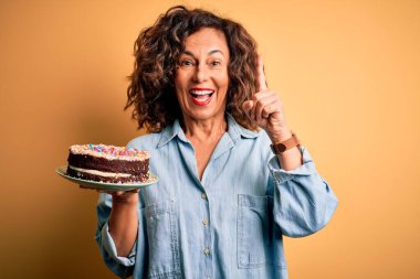 Orta yaşlı, güzel bir kadın, izole edilmiş sarı arka planda duran pastayı tutarken bir fikir ya da soruyla şaşırır.