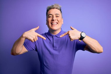 Soyutlanmış arka planda sıradan mor tişörtler giyen genç ve modern bir adam. Yüzünde bir gülümsemeyle, kendini gururlu ve mutlu bir şekilde gösteren bir adam..