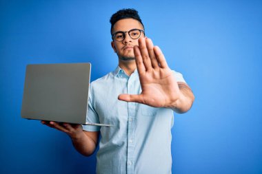 Gözlük takan genç yakışıklı iş adamı mavi arka planda dizüstü bilgisayar kullanıyor. Açık elle dur işareti yapıyor. Ciddi ve kendinden emin bir ifadeyle, savunma jesti yapıyor.