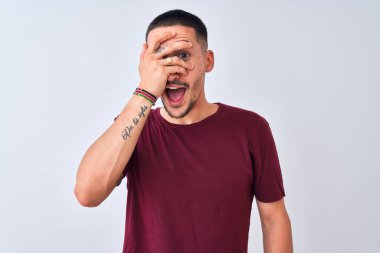 Young handsome man standing over isolated background peeking in shock covering face and eyes with hand, looking through fingers with embarrassed expression.