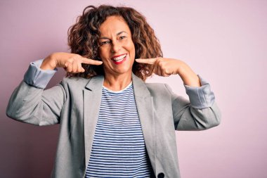 Soyutlanmış pembe arka planda zarif bir ceket giyen orta yaşlı, güzel bir iş kadını gülümseyen, parmaklarını ve ağzını gösteren. Diş sağlığı konsepti.